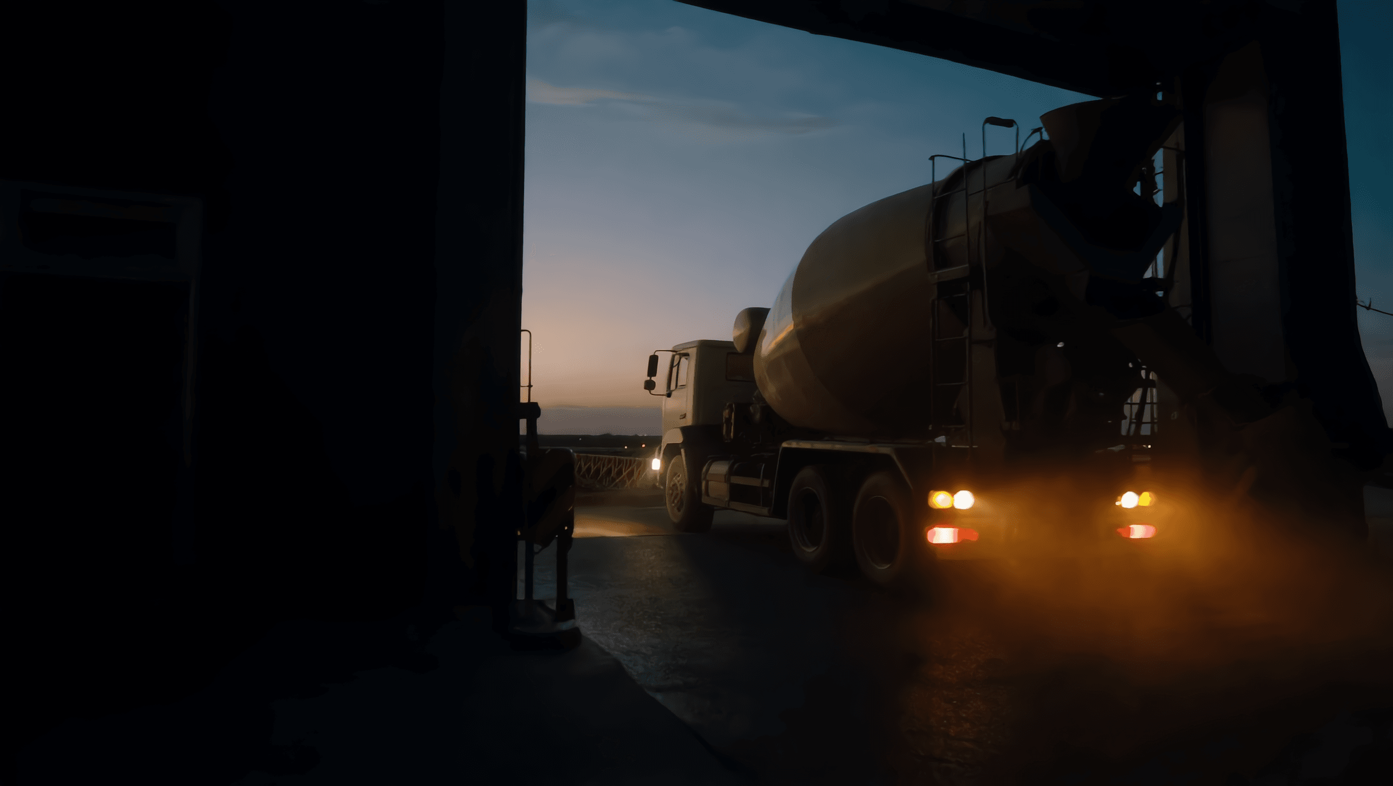 A concrete mixer truck driving out through a garage opening at dusk